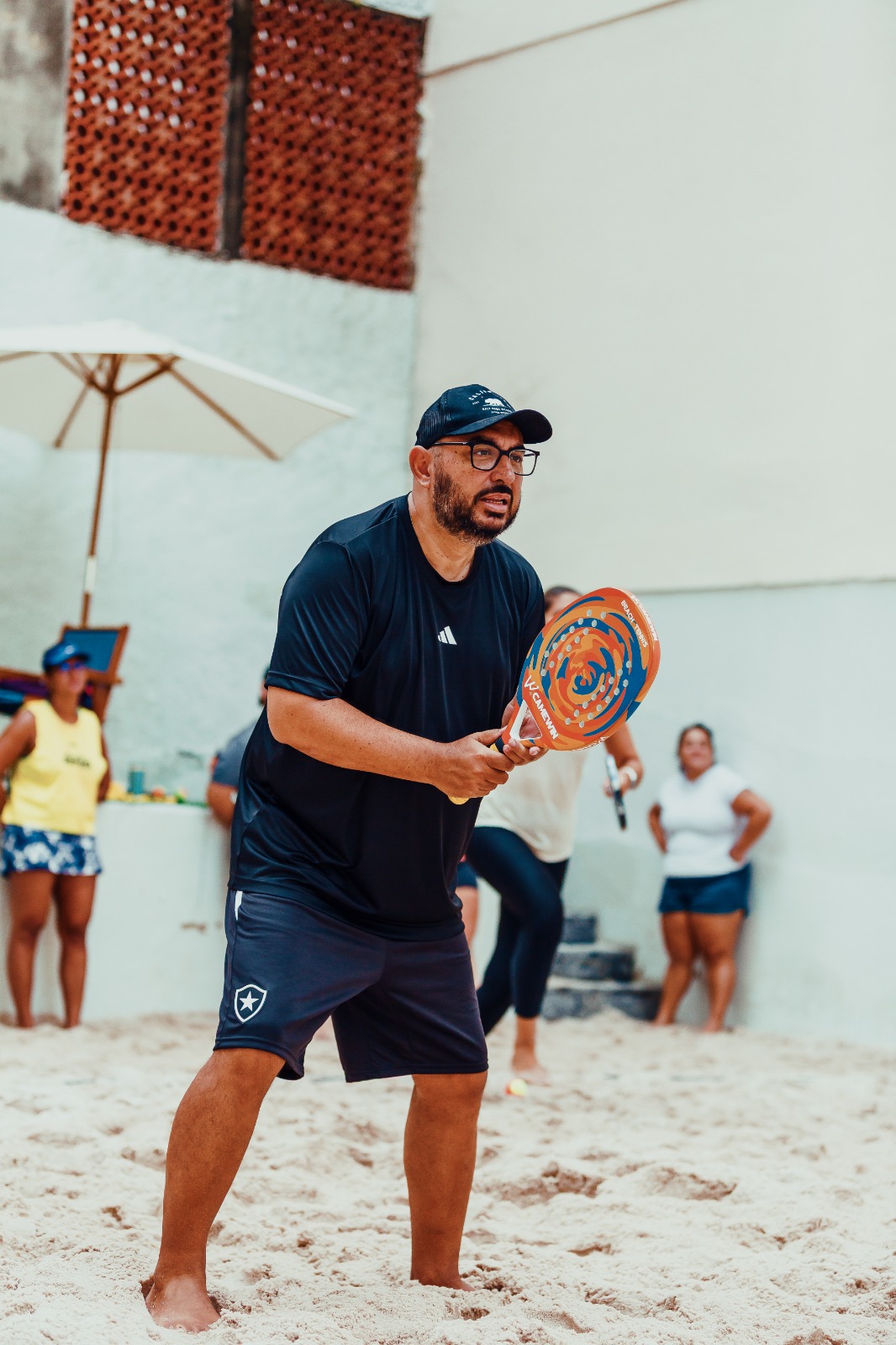 Jogador de boné, com a raquete pronta à frente do corpo, atento ao lance na quadra de areia da Arena Aldeia